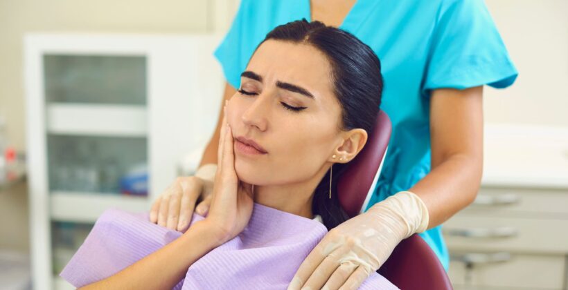 Young woman sitting in a dentist chair holding the side of her face with a faceless dentist behind her - Midland Bay Dental.